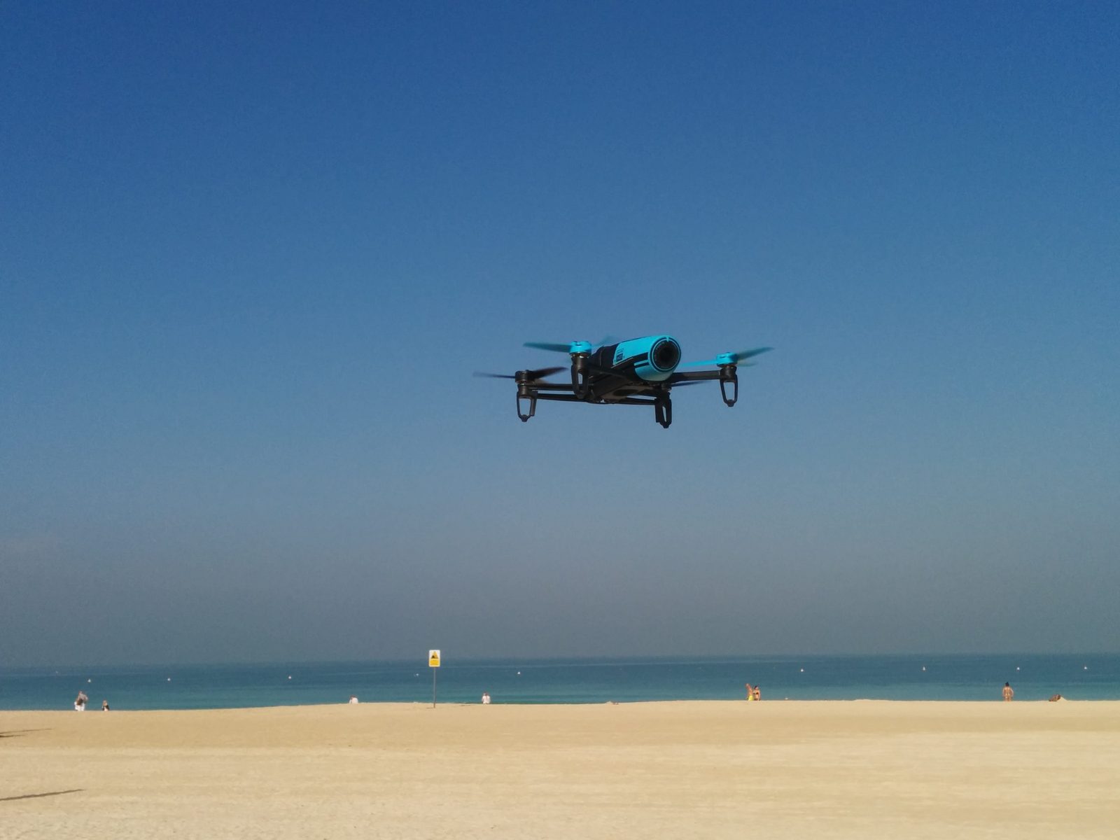 A small quadcopter drone flying over a Dubai beach under a clear blue sky.